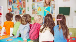 Teacher with a group of preschool children in a nursery. The children are sitting on the floor and listening teacher. Learning letters. In the background we can see a shelf with some, toys, black board and books. View from behind.