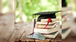 A graduation cap and diploma on a stack of books next to a face mask