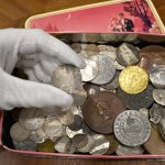 Image of the tin box where a rare coin from colonial Boston was found. A gloved hand holds the coin above the box.