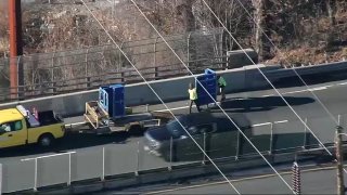 Large blue Amazon carts on wheels block the breakdown lane of a highway. DOT workers in neon vests are working to remove them with a pickup truck