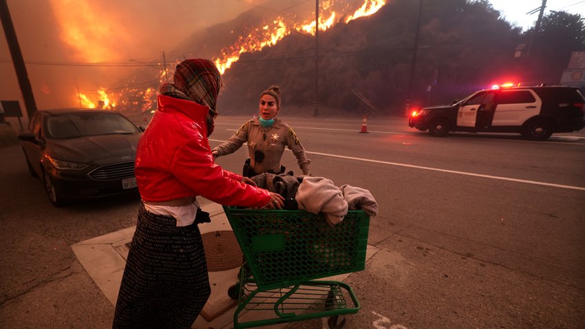 Pacific Palisades, CA – January 07: A police officer escorts a homeless woman to evacuate away from Pacific Coast Highway and Topanga Canyon Blvd as the Palisades Fire rages down the hills in Pacific Palisades, Calif. on Tuesday, Jan. 7, 2025. (Wally Skalij / Los Angeles Times via Getty Images)