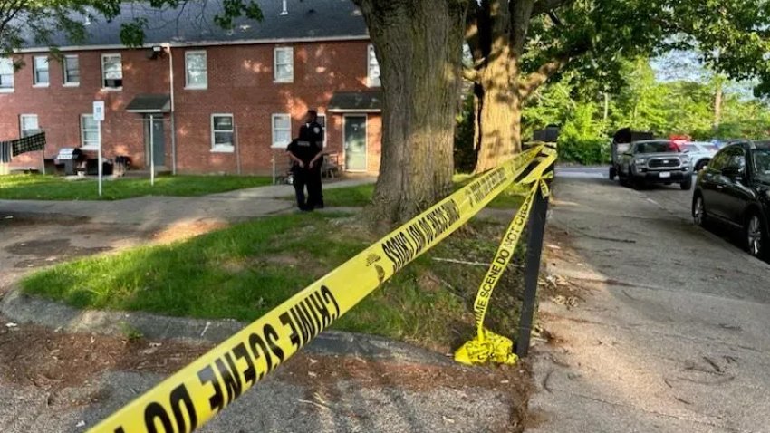 The scene of a deadly shooting in Providence, Rhode Island, on June 5, 2025. Brick buildings can be seen in the background, with police tape marking off an area of grass and trees in the foreground.